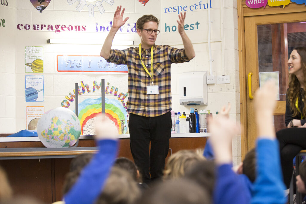 Artistic Director Brad McCormick stands in front of a group of young people in a school classroom with his arms in the air. 