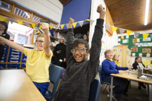 School students sitting at a desk with their hands raised in the air.