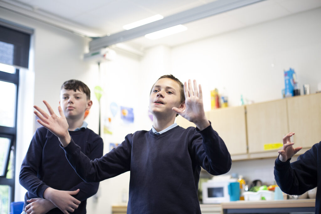 Two school students standing in a classroom, one has their hands lifted in front of them. 