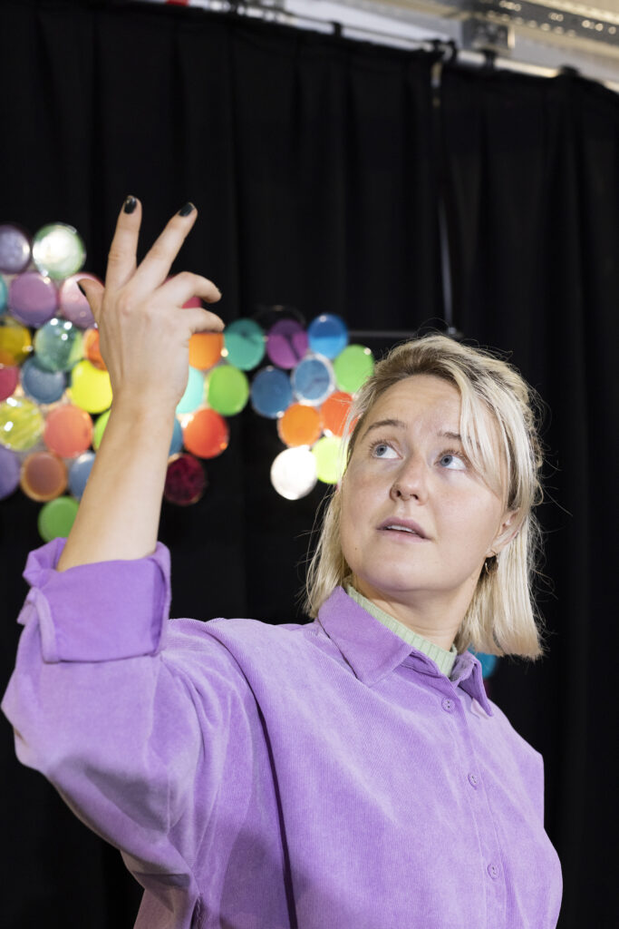 Jeannie raising her arm up and looking at her hand in front of a multi coloured set.