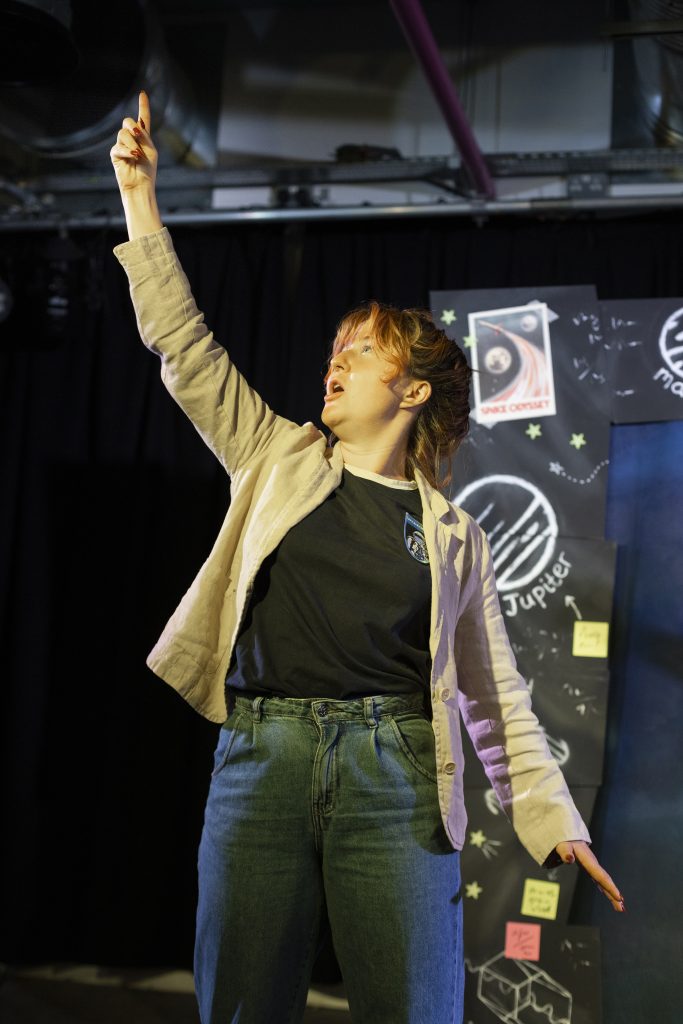 Jeannie pointing up into the sky in a post, in front of a rocket-themed stage set.