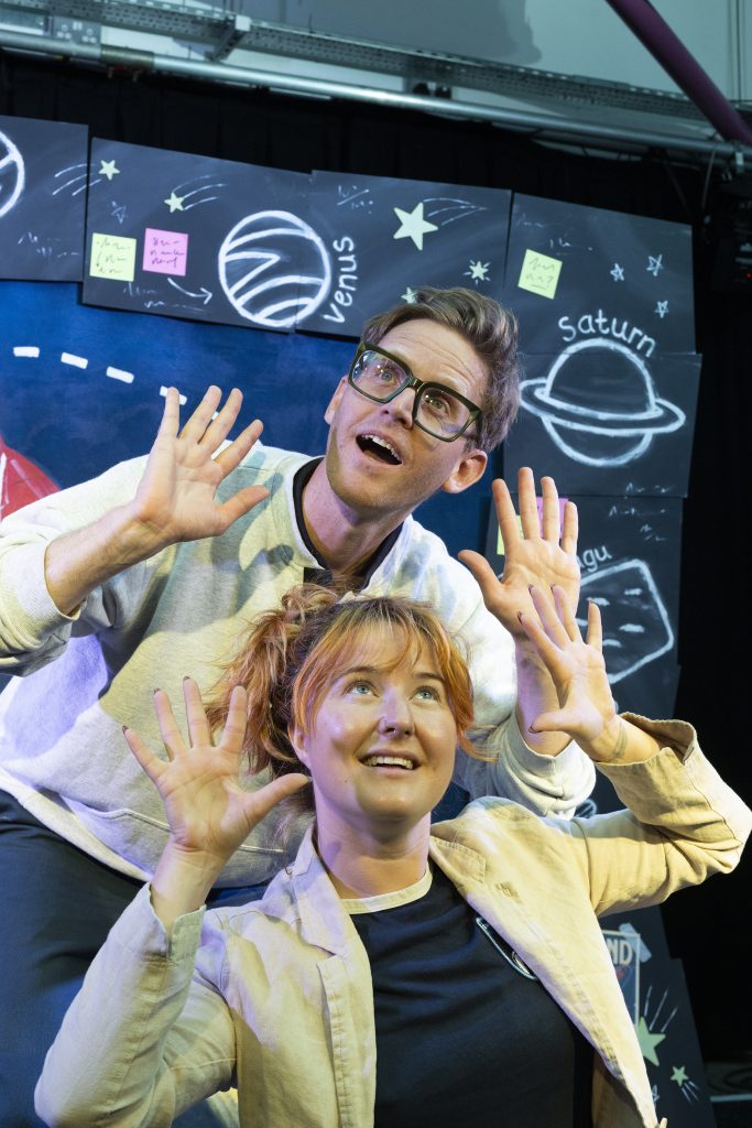 Jeannie and Brad both look up in wonder, hands raised beside their faces in a "wowed" pose, in front of the stage set.