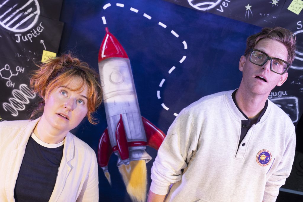 Jeannie and Brad, theatre facilitators, both gaze upward as if into space, standing in front of a rocket-themed stage set.