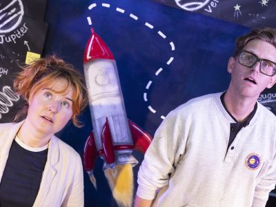 Jeannie and Brad, theatre facilitators, both gaze upward as if into space, standing in front of a rocket-themed stage set.