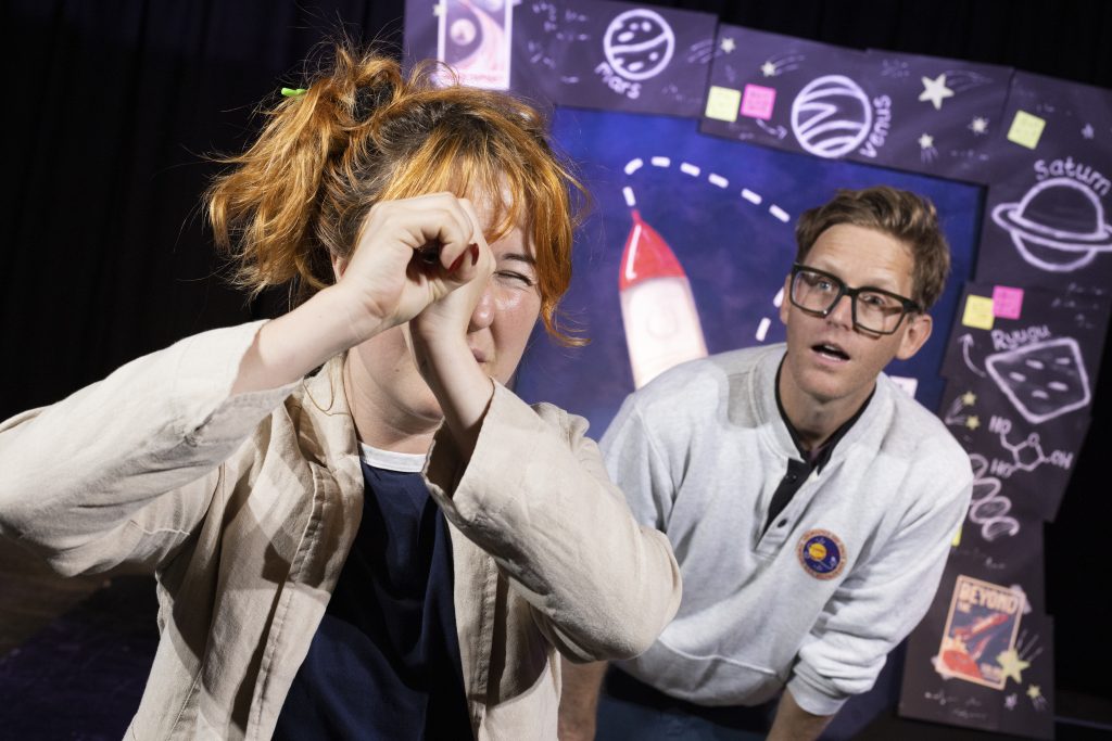 Jeannie peers through an imaginary telescope while Brad looks curiously behind him, both standing in front of the rocket stage set.