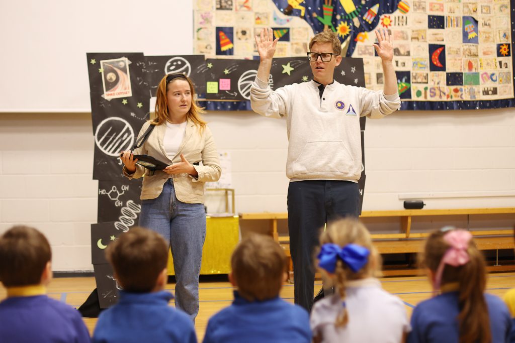 Jeannie and Brad stand in front of a class, leading a drama workshop in the school hall in front of a staged set.