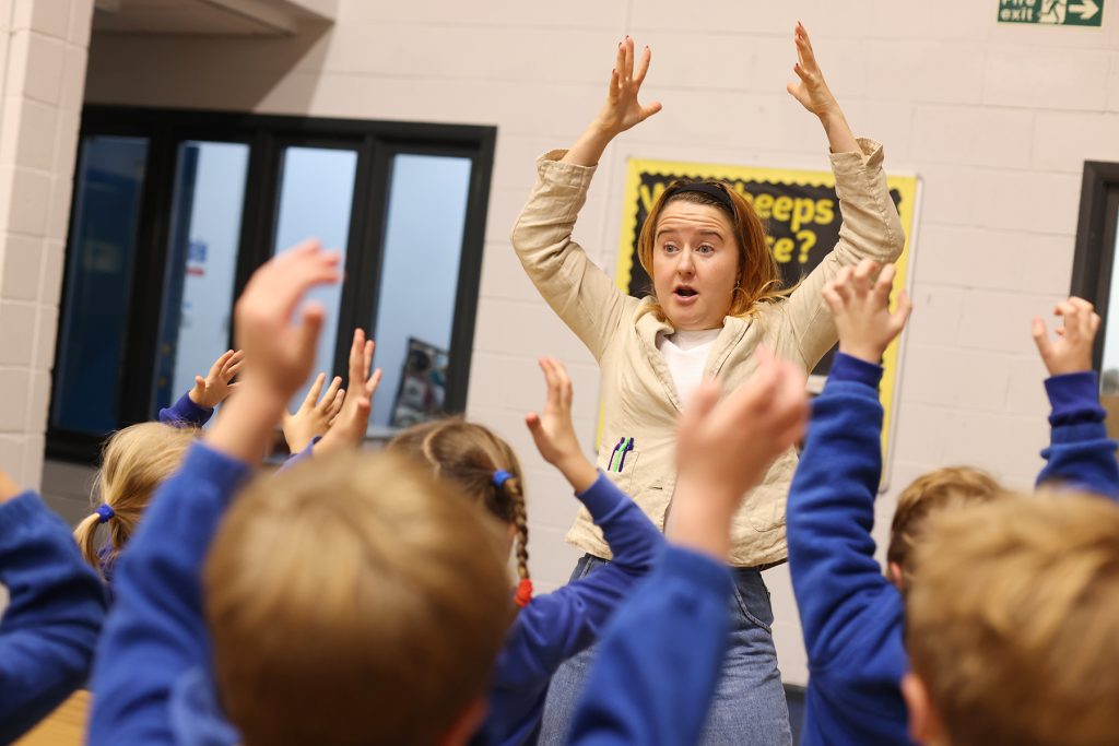 Jeannie facilitates a drama workshop with her hands raised as a group of children in front of her also raise their hands.