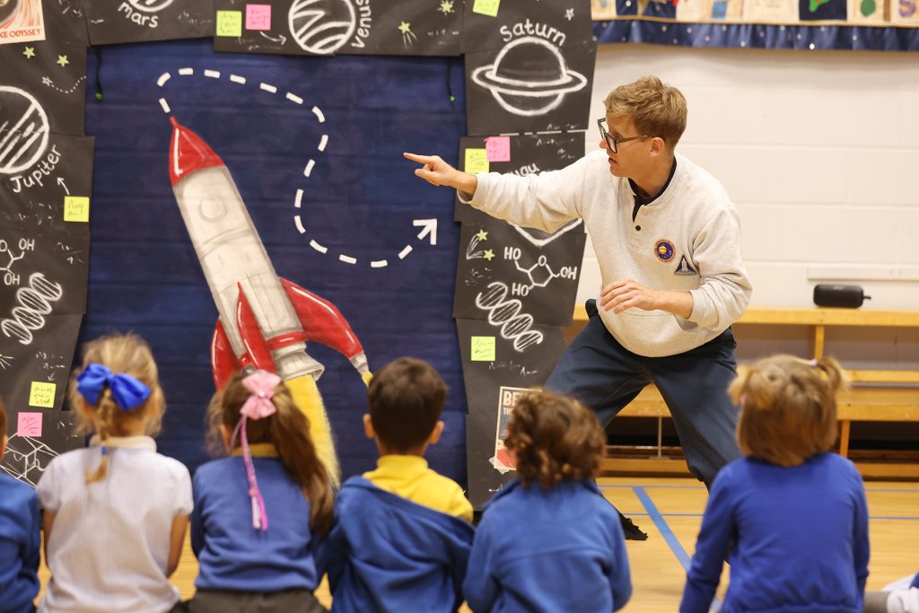 Brad points towards a rocket on the stage set in front of a group of children.