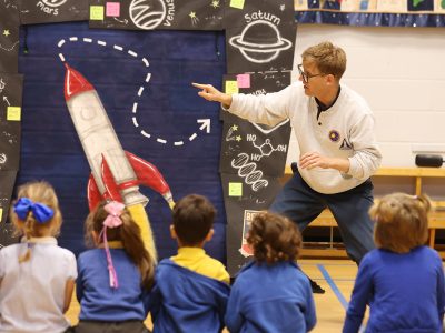 Brad points towards a rocket on the stage set in front of a group of children.